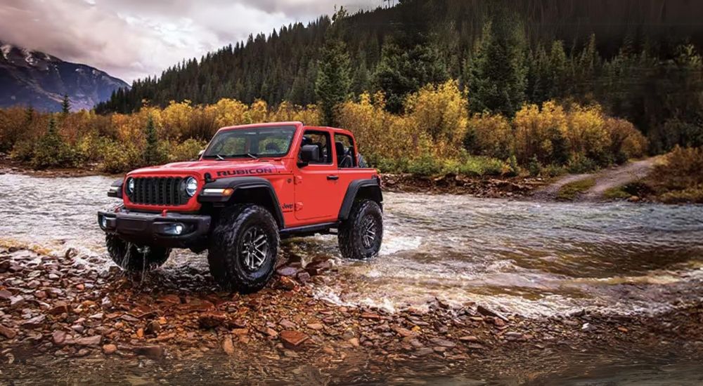 A red 2024 Jeep Wrangler Rubicon is shown driving thought a river after visiting a Jeep dealership near you.